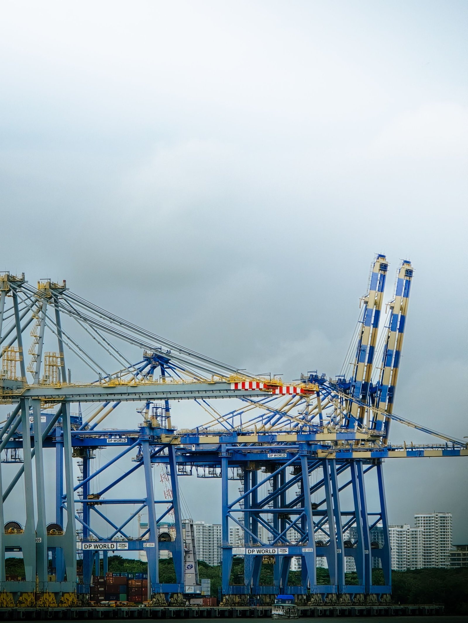 Blue and white cranes at a bustling port with shipping containers, under cloudy skies.